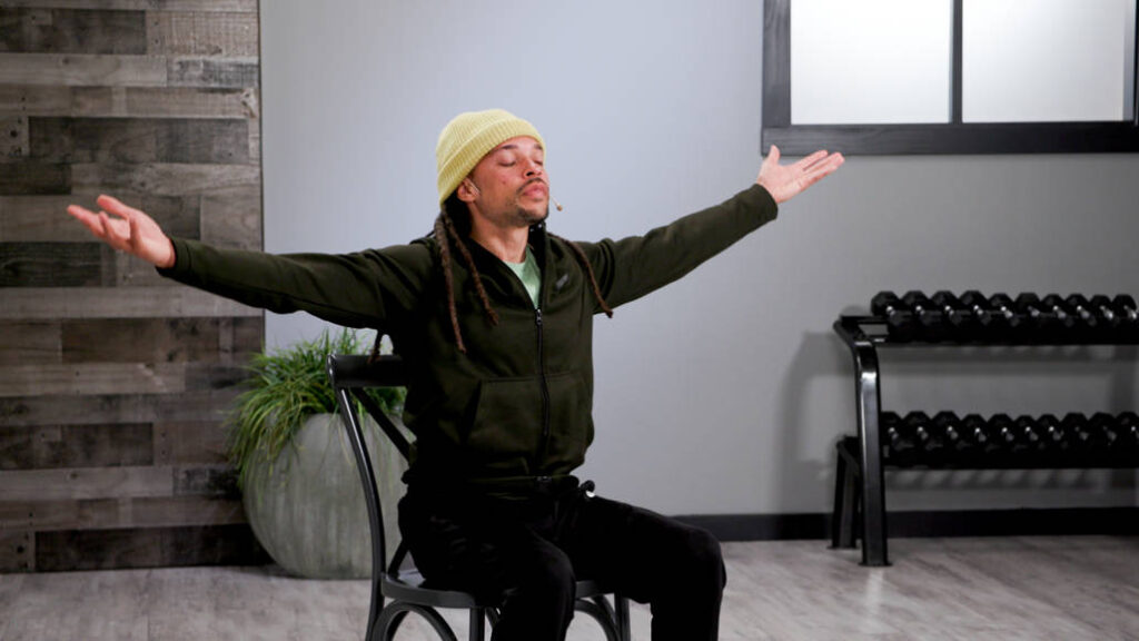 Wellbeats instructor Chance Y. is seated in a chair with his arms spread wide and his eyes closed leading a yoga session.