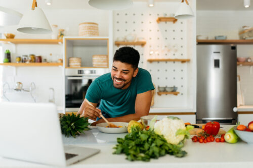 A man smiles while leaning on a kitchen counter surrounded by ingredients and a laptop.