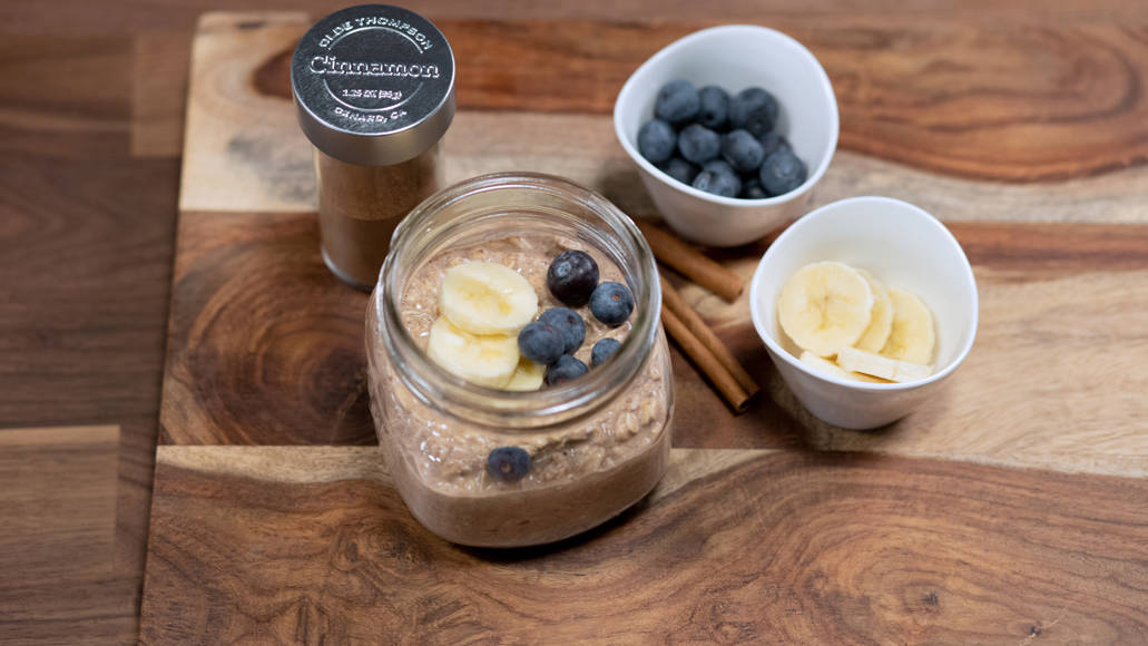 Overnight oats are pictured in a jar alongside bananas, blueberries, and cinnamon.